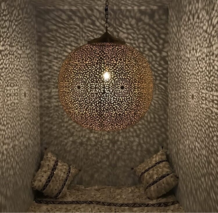 A brass ceiling light with intricate patterns hanging in a dimly lit space, creating a warm, ambient glow on the walls around it.